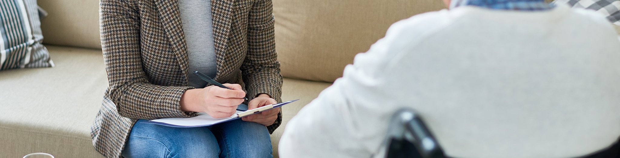 Das Bild zeigt eine Therapeutin mit einem Patienten im Rollstuhl. Sie schreibt sich etwas auf.