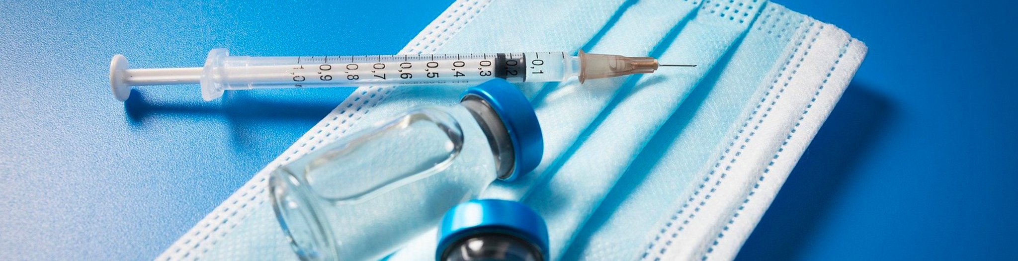 Das Bild zeigt eine Injektionsspritze und Impfdosen auf einer medizinischen Maske 