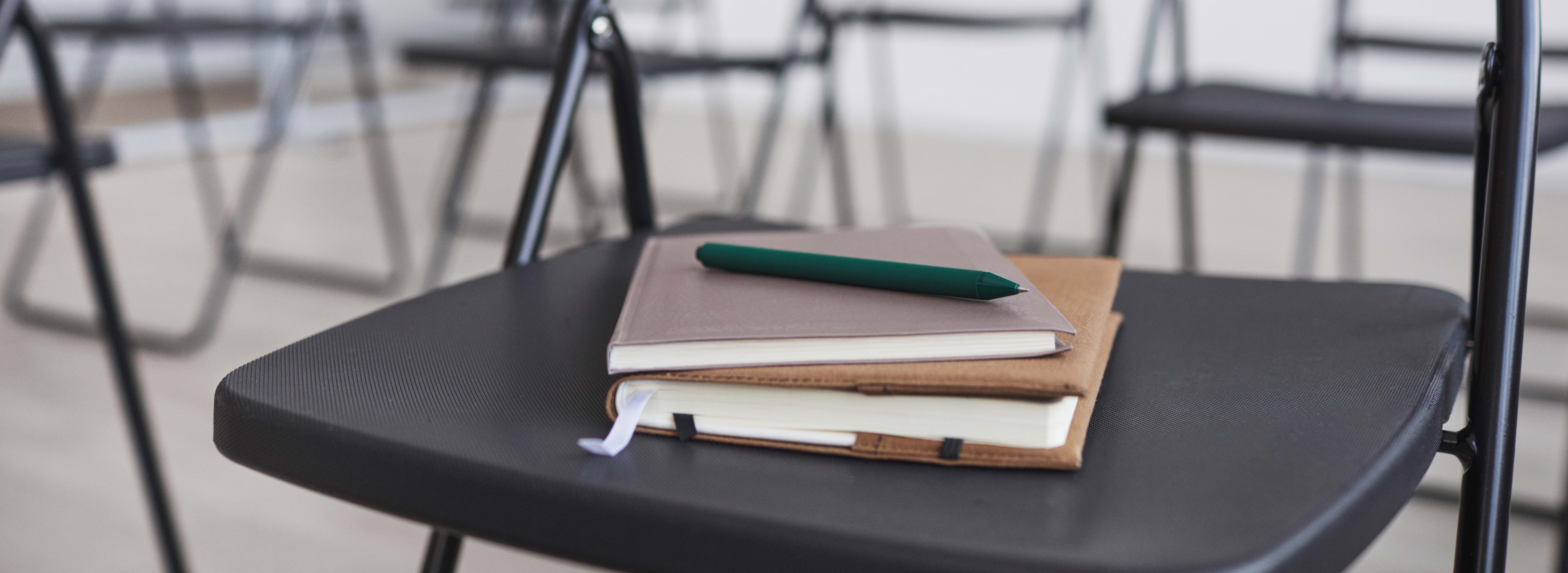 Das Foto zeigt einen Konferenzraum mit Stuhl im Vordergrund, mit Stift und zwei Notizbüchern auf der Sitzfläche