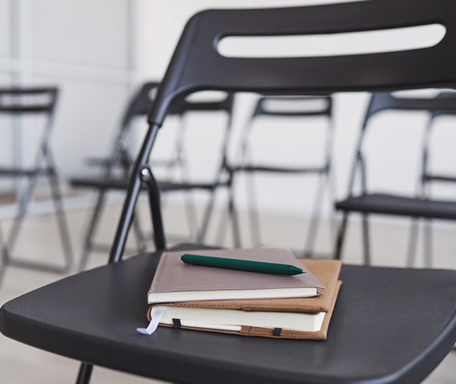 Das Foto zeigt einen Konferenzraum mit Stuhl im Vordergrund, mit zwei Notizbüchern und einem Stift auf der Sitzfläche